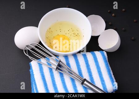 Zerbrochenes Hühnerei mit schwarzem Pfeffer. Es ist bereit, mit Whisker behackt zu werden. Es ist auf schwarzem Hintergrund mit blau gestreiften Servietten gehalten. Stockfoto