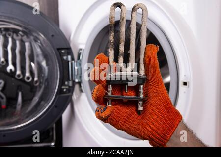 Reparatur von Waschmaschinen. Hand eines Reparaturmanns mit einem turbulenten elektrischen Heizgerät, das mit einer Beschichtung aus hartem Wasser bedeckt ist. Austausch des elektrischen Heizgeräts Stockfoto