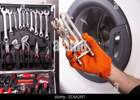 Reparatur von Waschmaschinen. Hand eines Reparaturmanns mit einem turbulenten elektrischen Heizgerät, das mit einer Beschichtung aus hartem Wasser bedeckt ist. Austausch des elektrischen Heizgeräts Stockfoto