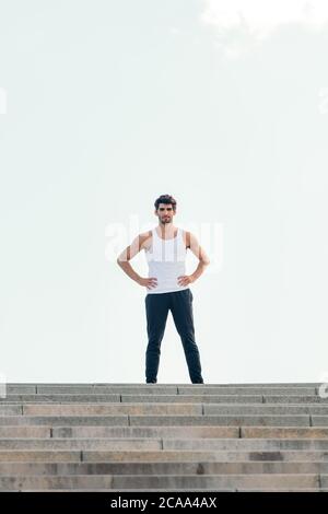 Vertikales Foto eines gutaussehenden Sportlers, der mit den Händen auf der Hüfte oben auf einer Treppe steht, Konzept des urbanen Sports und des gesunden Lebensstils, Co Stockfoto