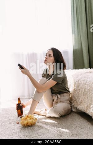 Eine junge Frau trinkt Bier und isst Chips zu Hause. Entspannen Sie sich zu Hause beim Fernsehen und Filmen. Stockfoto