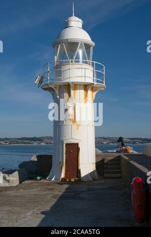 Blick auf den Hafen von Brixham Rosting Lighthouse am Ende des Hafenpiers Blick auf die ganze Struktur Mann sitzt auf einem Stuhl und schaut hinaus Blue Sky Hochformat Stockfoto