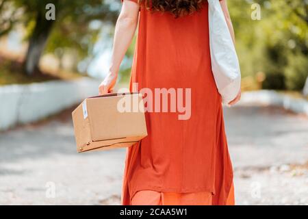 Eine Frau in einem roten Kleid geht die Straße entlang mit einem Paket in der Hand. Rückansicht. Das Konzept der Lieferung von Waren. Stockfoto