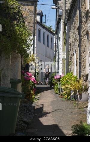 Sonniger Tag in Mousehole enge Straße Person zu Fuß entfernt Old Gebäude auf beiden Seiten Blumen und Sträucher in der Straße Sonnenschein Blue Sky Hochformat Stockfoto