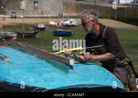 Sonniger Tag im Hafen von Mousehole. Mann, der an einem umgedrehten Rumpf eines Bootes arbeitet. Mit Dichtungspistole. Boote in der Ferne aus dem Fokus. Querformat Stockfoto