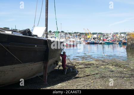 Newlyn Old Harbour Traditional Lugger wird restauriert man Reinigungsseite Von Rumpf Nahaufnahme des Schiffes mit Hafenbooten Und Trawler im Hintergrund Stockfoto