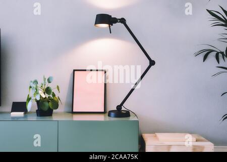 Stilvolles modernes Interieur, leerer Bilderrahmen mit schwarzer Lampe und grüner chinesischer Geldanlage, Retro-Pfannkuchen-Pflanze auf grünem Tisch skandinavisches Design Stockfoto