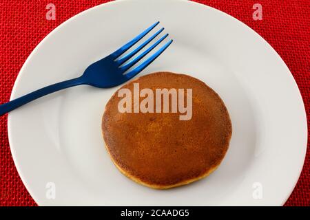 Kleines Pfannkuchen-Sandwich mit Himbeermarmelade-Füllung auf weißem Snackteller mit blauer Gabel auf roter Tischdecke Stockfoto