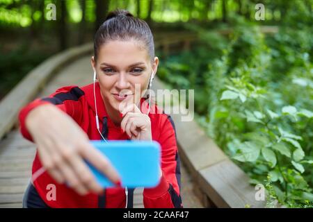 Konzept des Fernlernens. Ein attraktives Mädchen in Sportbekleidung sitzt auf einem Holzweg in einem Waldpark. Online-Unterricht per Smartphone. Stockfoto