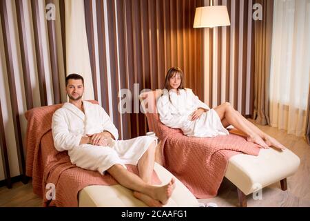 Zwei junge Menschen in bequemen Bademänteln, die auf den Sesseln ruhen. Seitenansicht full lenngth Foto. Stockfoto
