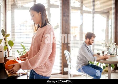 Junges Ehepaar in der Küche. Eine Brünette sitzt auf einem Stuhl und benutzt ein digitales Tablet. Ein Mann, der neben dem Tisch steht und eine Tasse Kaffee hält. Stockfoto