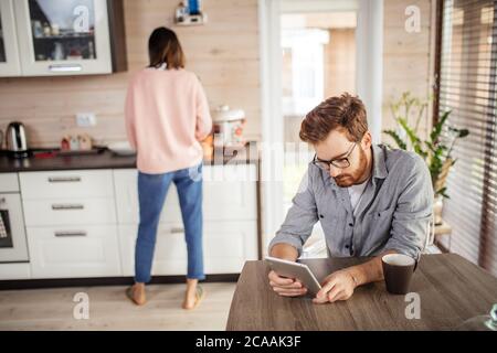 Junges Ehepaar in der Küche. Eine Brünette sitzt auf einem Stuhl und benutzt ein digitales Tablet. Ein Mann, der neben dem Tisch steht und eine Tasse Kaffee hält. Stockfoto