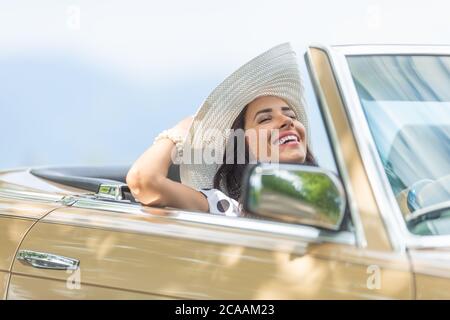 Rechtshändige Fahrerin mit geschlossenen Augen, Kopf nach hinten gekippt hält ihren Hut und genießt den Sommer-Cabriolet-Antrieb. Stockfoto