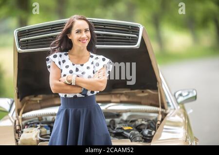 Selbstbewusste Frau steht vor einem Auto mit offener Motorhaube, Arme gekreuzt, lächelnd in die Kamera. Stockfoto