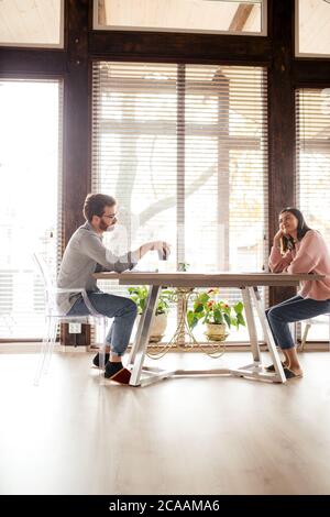 Panoramablick auf das Paar, das sich gegenüber am Tisch in einem großen, luftigen Raum sitzt und nach dem Mittagessen zu Hause über Pläne diskutiert Stockfoto