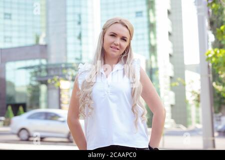 Frau mit langen blonden Haaren trägt weißes elegantes Hemd und steht auf der Straße Blick auf die Kamera Stockfoto