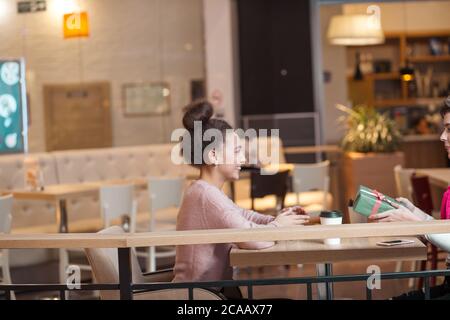 Brünette Frau Geschenke Geschenk, um ihre blonde Freundin während der Sitzung in Mall Cafe. Das blonde Mädchen ist auf der Suche glücklich und überrascht. Shopping, Menschen Stockfoto