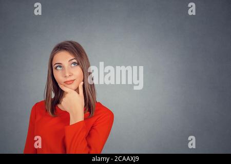 Nachdenkliche Frau in rotem Kleid. Portrait glücklich schöne Frau denken Blick nach oben isoliert graue Wand Hintergrund mit Kopieplatz. Gesichtsausdruck des Menschen Stockfoto