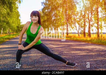 Junge Frau, die sich die Beine vor dem Joggen auf Asphaltstraße in der Landschaft streckt Stockfoto