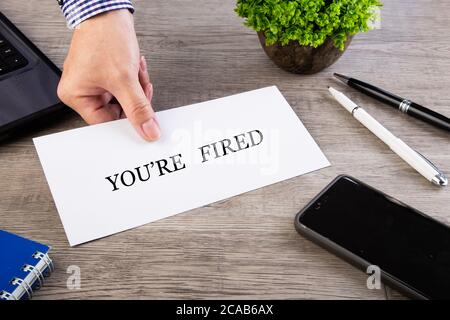 Nahaufnahme einer Hand, die ein Papierschild mit hält Text [YOU'RE FIRED] auf einem Büroschreibtisch Stockfoto