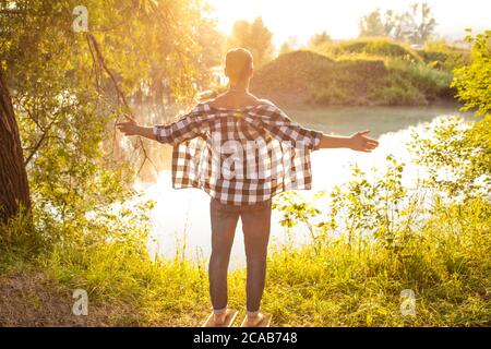 Junger Mann mit T-Shirt und Jeans auf dem Holzbrett mit Metallnägeln am Ufer des Flusses und Stretching Arme stehen. Rückansicht Foto. Heilen Stockfoto