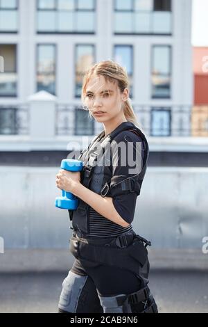 Blonde Frau in elektrische Muskelstimulation Anzug stehen mit blauen Hantel. Nahaufnahme. Stockfoto