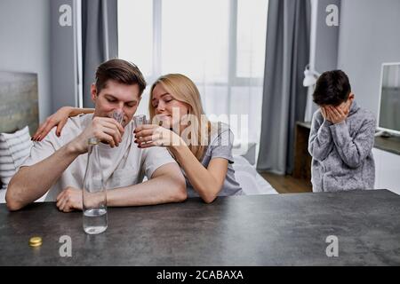 Das Kind braucht Liebe, aber seine Eltern kümmern sich nicht, sitzen sie zusammen Alkohol trinken. Kind Junge weinen im Hintergrund. Drinnen, zu Hause Stockfoto