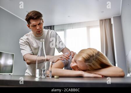 Der junge Kaukasier lebt mit einer wertlosen Alkoholikerin, er ist des Lebens mit ihr müde, die Frau liegt bewusstlos auf dem Tisch Stockfoto