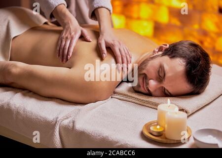 Der junge Kaukasusmann entspannt sich während einer Massage auf dem Rücken, die von weiblichen Händen gemacht wurde, auf dem Spa-Tisch liegend und genießt eine Spa-Behandlung Stockfoto