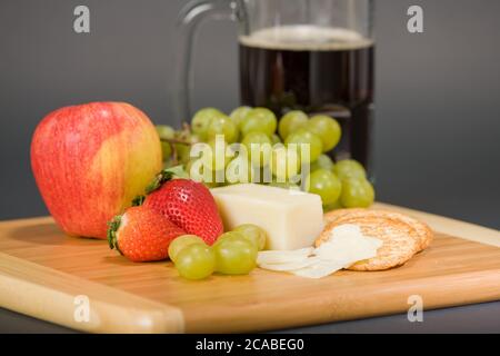 Gruyere Schweizer Käse, Becher Bier, Obst und Vollkorncracker auf einem hölzernen Schneidebrett Stockfoto