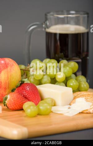 Gruyere Schweizer Käse, Becher Bier, Obst und Vollkorncracker auf einem hölzernen Schneidebrett Stockfoto