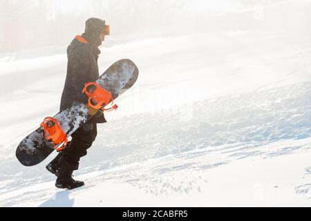 Nachdenklicher Kerl läuft mit einem Snowboard auf einem Hügel. Copy Space. Motivierter Mann versucht, die Spitze des Berges an einem frostigen Tag zu erreichen Stockfoto