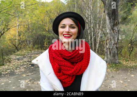 Schöne junge Frau in einem weißen Mantel, gehäkeltem rotem Schal und schwarzem Hut. Lächelndes Porträt im Freien Stockfoto
