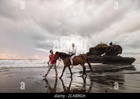 Asien, Indonesien, Bali, junger kaukasischer Mann mit einer schönen jungen kaukasischen Frau, die auf einem Pferd sitzt, am Strand bei Sonnenuntergang, mit typisch balinesischem Felsen Stockfoto