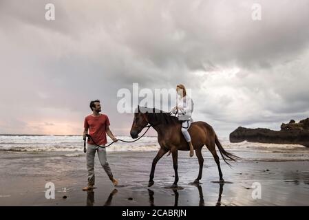 Asien, Indonesien, Bali, junger kaukasischer Mann mit einer schönen jungen kaukasischen Frau, die auf einem Pferd sitzt, am Strand bei Sonnenuntergang, mit typisch balinesischem Felsen Stockfoto