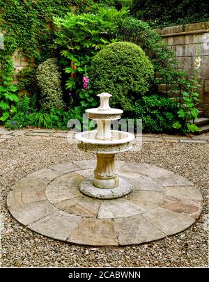 Ein Stein gemauerter Cottage Garten eine zentrale dreistufige Steinwasseranlage und reife Sträucher und Hollyhocks. Stockfoto