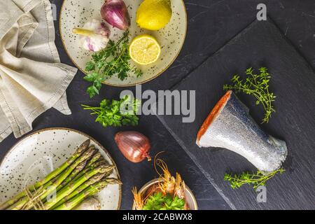 Forellen umgeben Petersilie, Zitrone, Garnelen, Garnelen, Spargel in Keramikplatten. Tischoberfläche aus schwarzem Beton. Gesunde Meeresfrüchte Hintergrund. Stockfoto