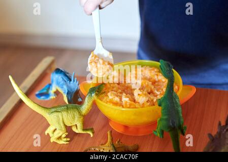 Spielzeug-Dinosaurier stehen auf einem Kindertisch neben einem Teller Haferbrei mit Löffel. Stockfoto