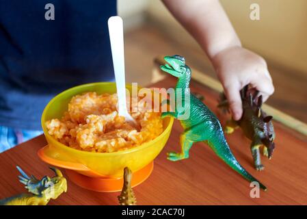 Spielzeug-Dinosaurier stehen auf einem Kindertisch neben einem Teller Haferbrei mit Löffel. Stockfoto