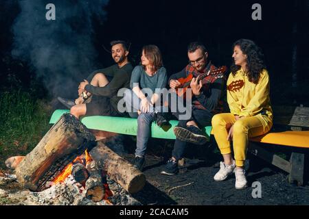 Vier Erwachsene Freunde sitzen auf einer bunten Bank im Wald, spielen Gitarre und singen. Vorderansicht einer Gruppe von Menschen, die die Wärme des brennenden Feuers in der Sommernacht genießen. Konzept des Tourismus, Camping. Stockfoto