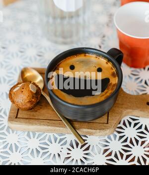 Draufsicht Smile Face Creama heißen schwarzen Kaffee in schwarzer Keramik Tasse auf Holztablett setzen weißen Tisch mit roten Plastikbecher und Glasflasche Wasser auf wh Stockfoto