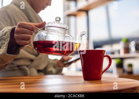 Frau mit Handy, während sie sich einen Tee serviert Stockfoto