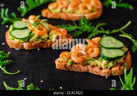 Leckeren snack Toast mit Avocado, gebratene Garnelen, Gurke und Rucola auf schwarzem Hintergrund. Selektiver Fokus, Nahaufnahme Stockfoto