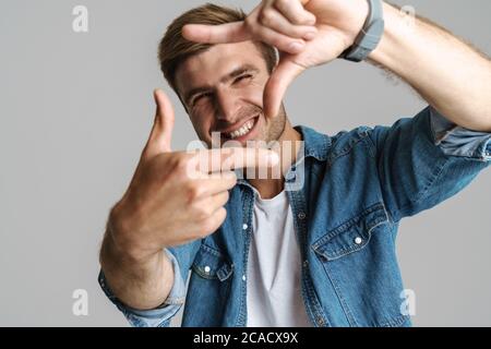 Portrait von fröhlich unrasiert Mann lächeln und machen Frame-Geste auf Kamera isoliert auf grauem Hintergrund Stockfoto