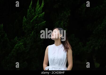 Ein asiatisches Mädchen in reinem weißen Rock, vor Kiefern unter natürlichem Tageslicht stehend. Unschärfem dunklen Hintergrund. Natürliche Schönheit Stockfoto