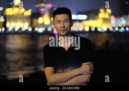 Ein asiatischer junger Mann, der nachts im Bund in Shanghai steht. Bunte defokussed Stadt Lichter Hintergrund Stockfoto