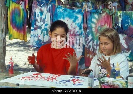 Hispanic- und Anglo-Mädchen malen vor gebürsteten T-Shirts, die an der Wäscheleine hängen, im Girl Scout Sommercamp am Lake Travis, Texas, USA. ©Bob Daemmrich Stockfoto