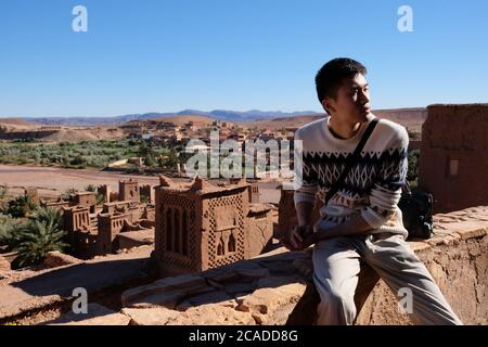 Ein asiatischer junger Mann, der auf der alten braunen Stadtmauer von Ait Ben Haddou unter Sonnenschein sitzt. Berühmte alte Berber kasbah in Marokko Stockfoto