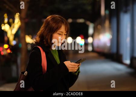 Eine asiatische junge Frau SMS-Nachricht auf Telefon draußen in der Nacht. Verwischen Sie farbenfrohe Straßenbeleuchtung im Hintergrund Stockfoto