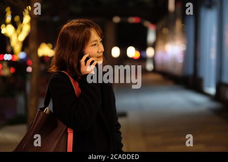 Eine asiatische junge Frau, die telefoniert und abends im Freien lächelt. Verwischen Sie farbenfrohe Straßenbeleuchtung im Hintergrund Stockfoto